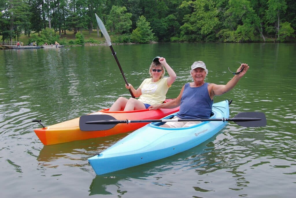 Two people waving from kayaks on a sunny lake surrounded by trees