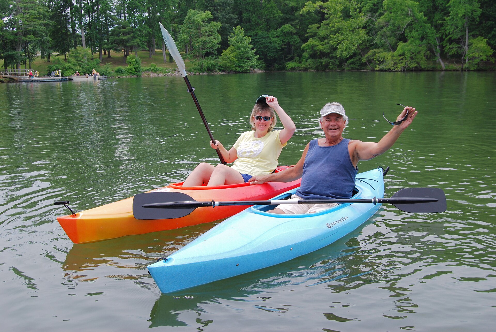 Two people waving from kayaks on a sunny lake surrounded by trees